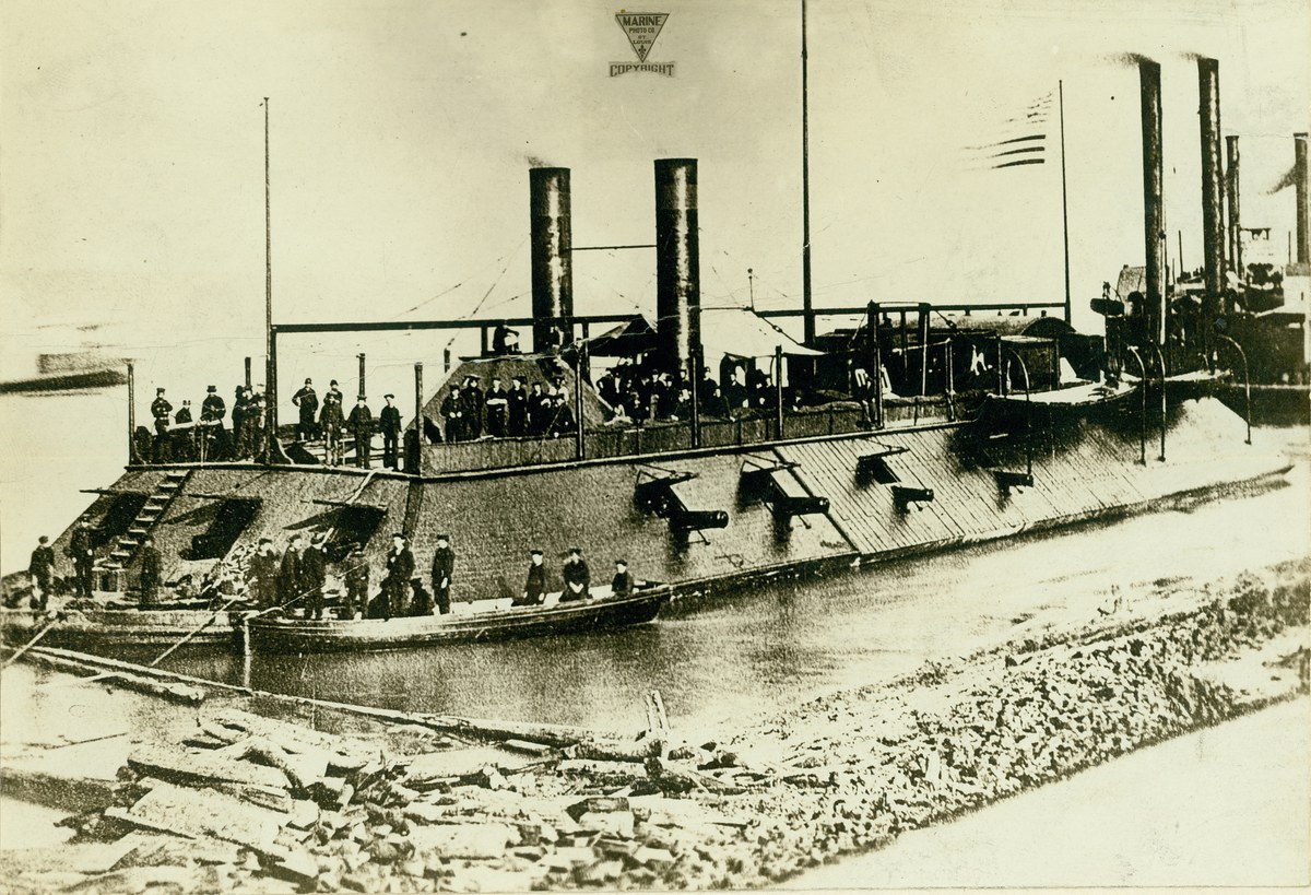 Photograph of an ironclad gunboat with sailors onboard. Two smaller rowboats are in the foreground with more sailors in them. In the background are ot, 1863